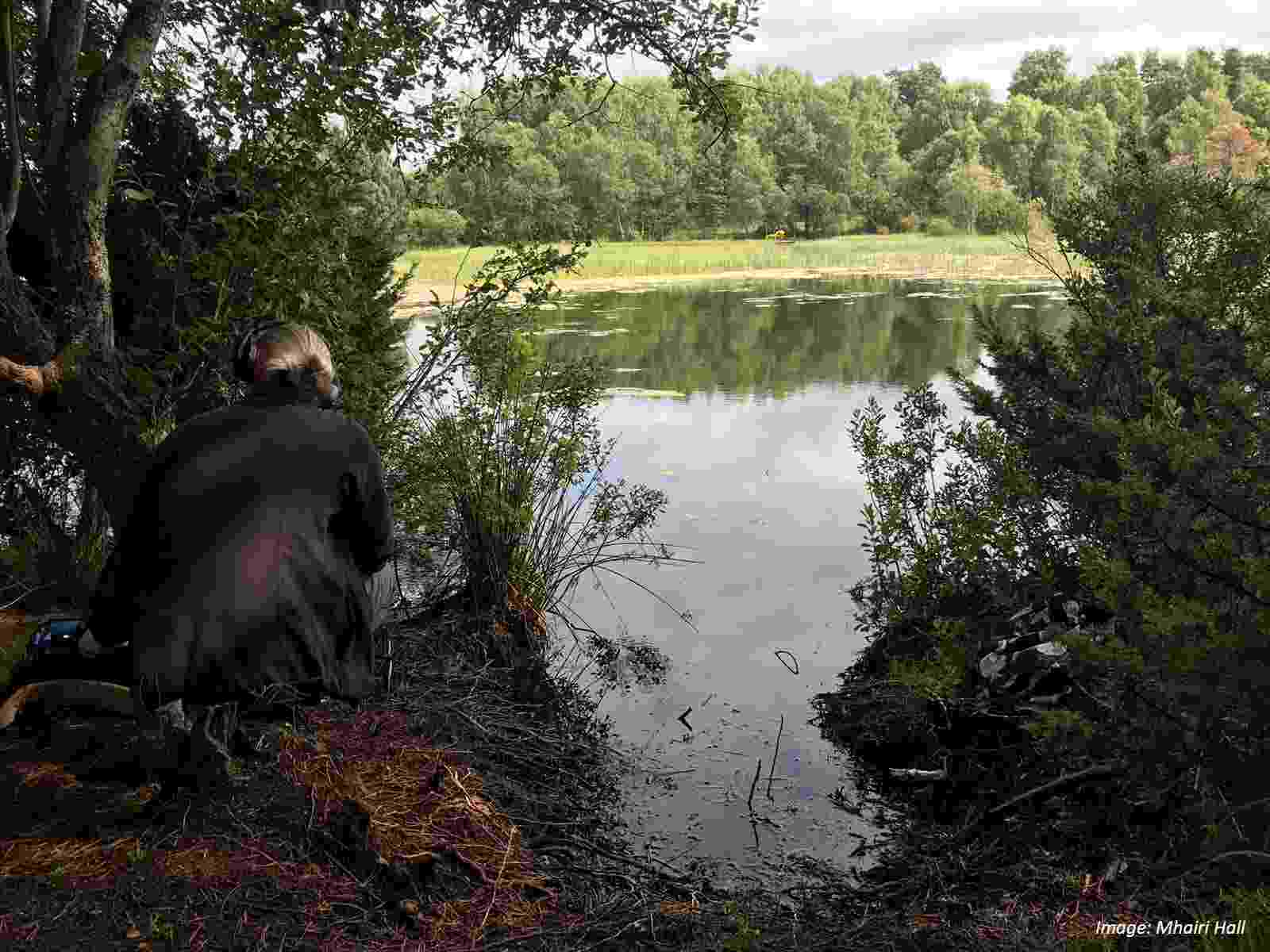 A photograph of the author crouching beside a lake, using audio equipment